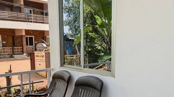 A balcony with two plastic chairs, a window with a view of greenery, and a sign indicating rooms are available.