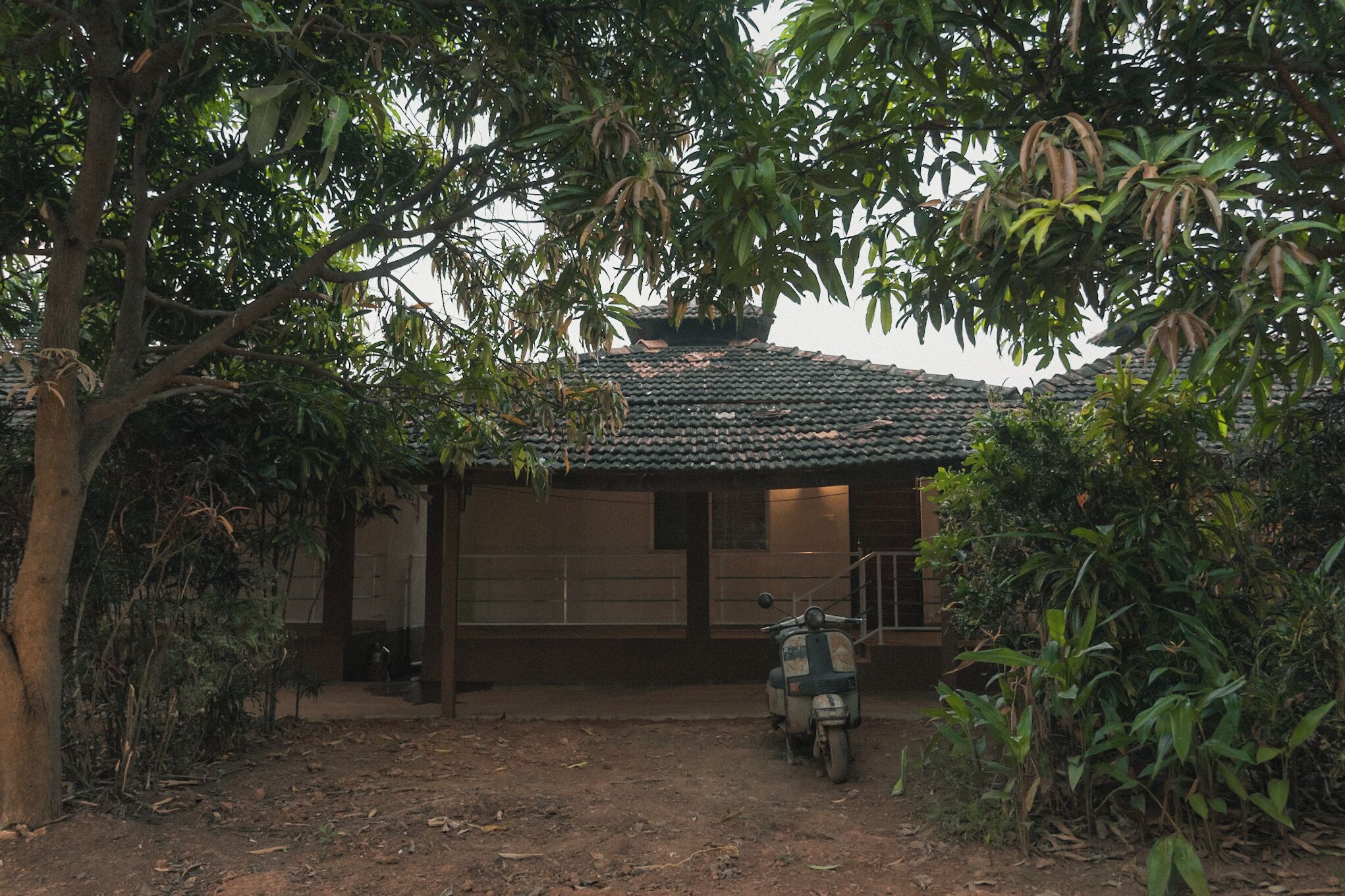 A rustic building with a tiled roof, surrounded by dense foliage and a dirt path leading to a parked scooter.