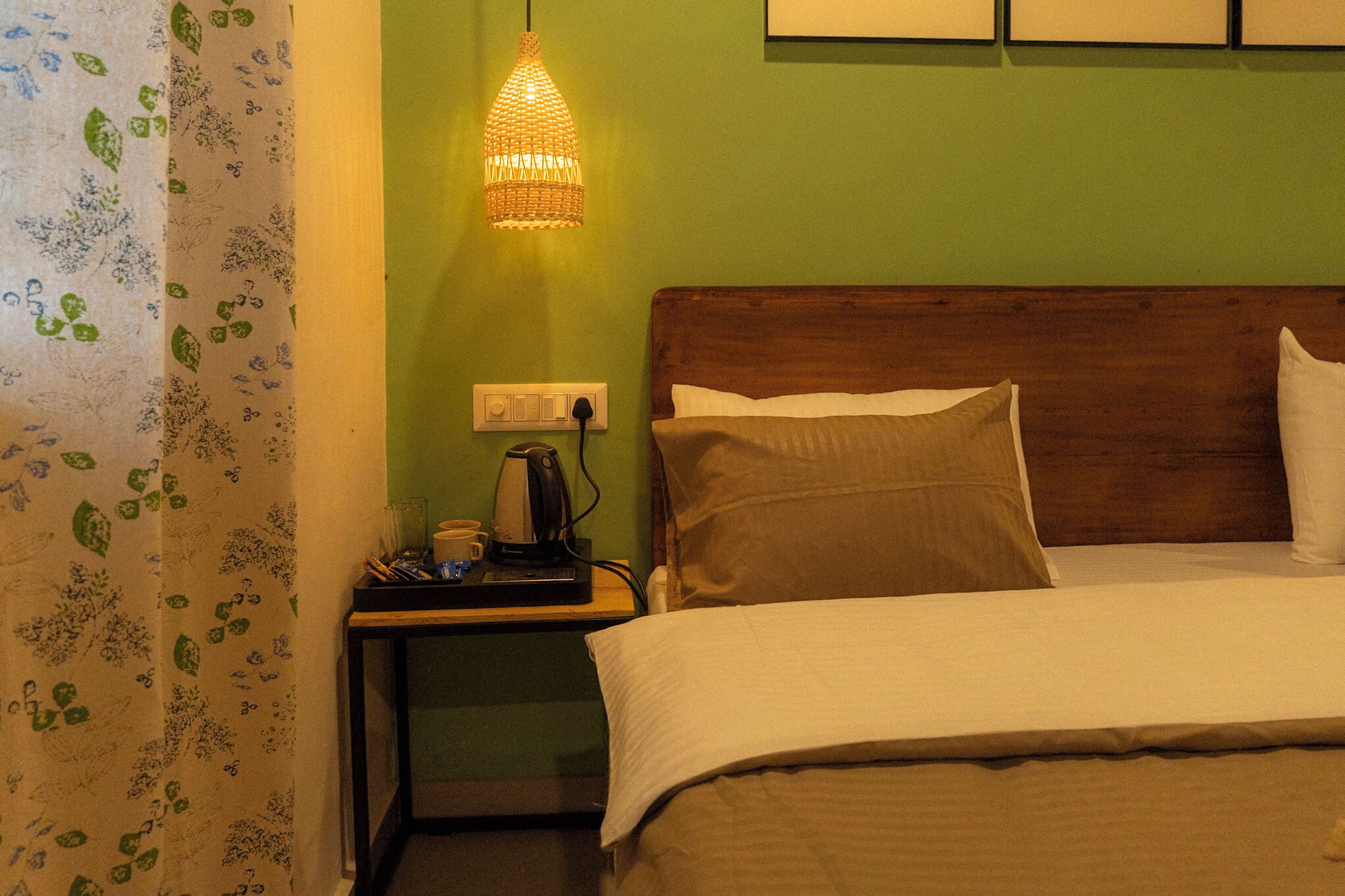 A hotel room with a bed, a nightstand with a kettle, and a wall-mounted light fixture.