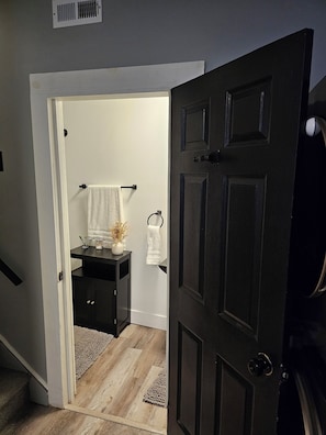 A doorway leading to a bathroom with a dark door, a white towel hanging on a rack, and a small vanity with a mirror and a vase with dried flowers. - Chic, Clean, Comfortable Luxury for all (Cleveland)