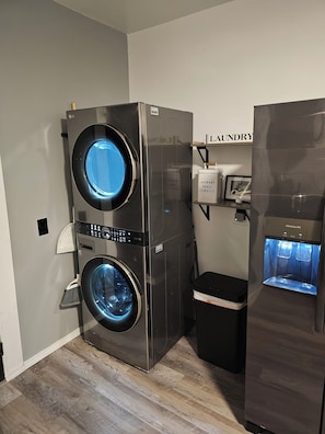 A laundry room with a stacked washer and dryer, a water dispenser, and a shelf with laundry supplies. - Chic, Clean, Comfortable Luxury for all (Cleveland)