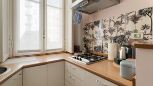 Een keuken met een gasfornuis, een raam met zonwering, een wand met behang van tropische planten en een kleine prullenbak.