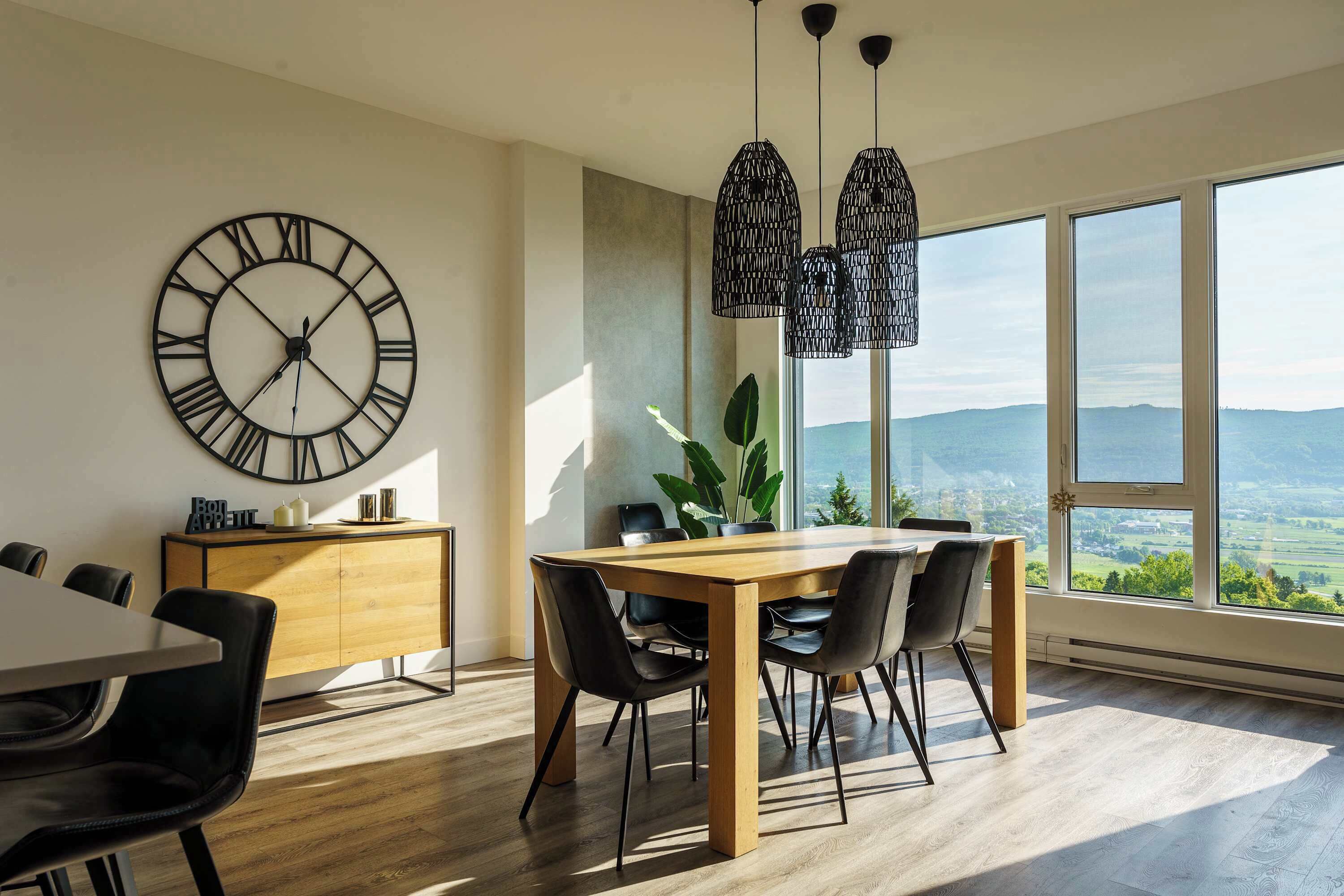Een eetruimte met een houten tafel, zwarte stoelen, een grote wandklok, hanglampen en een uitzicht door grote ramen.