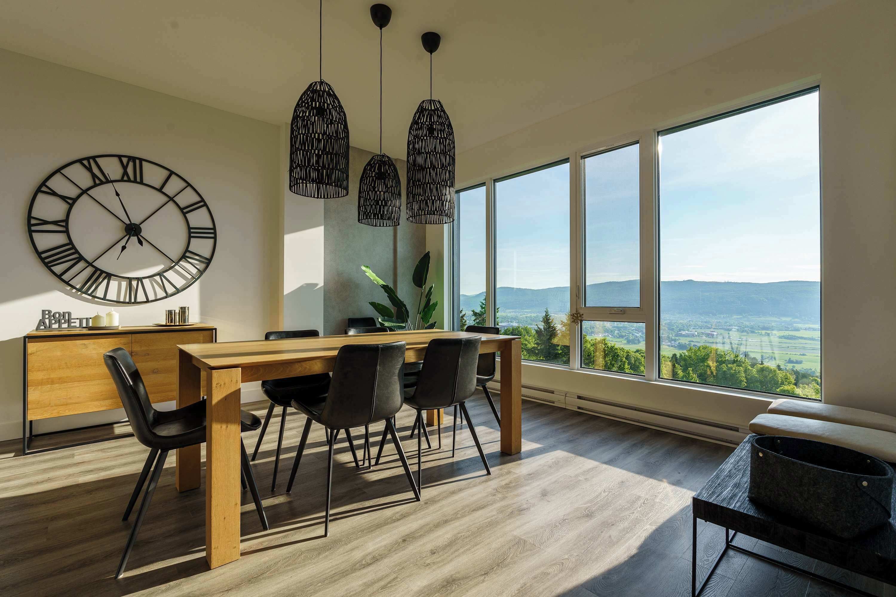 Un espace repas moderne avec une table en bois, des chaises noires, une grande horloge murale, des luminaires suspendus et une vue dégagée sur l’extérieur grâce à de grandes fenêtres.