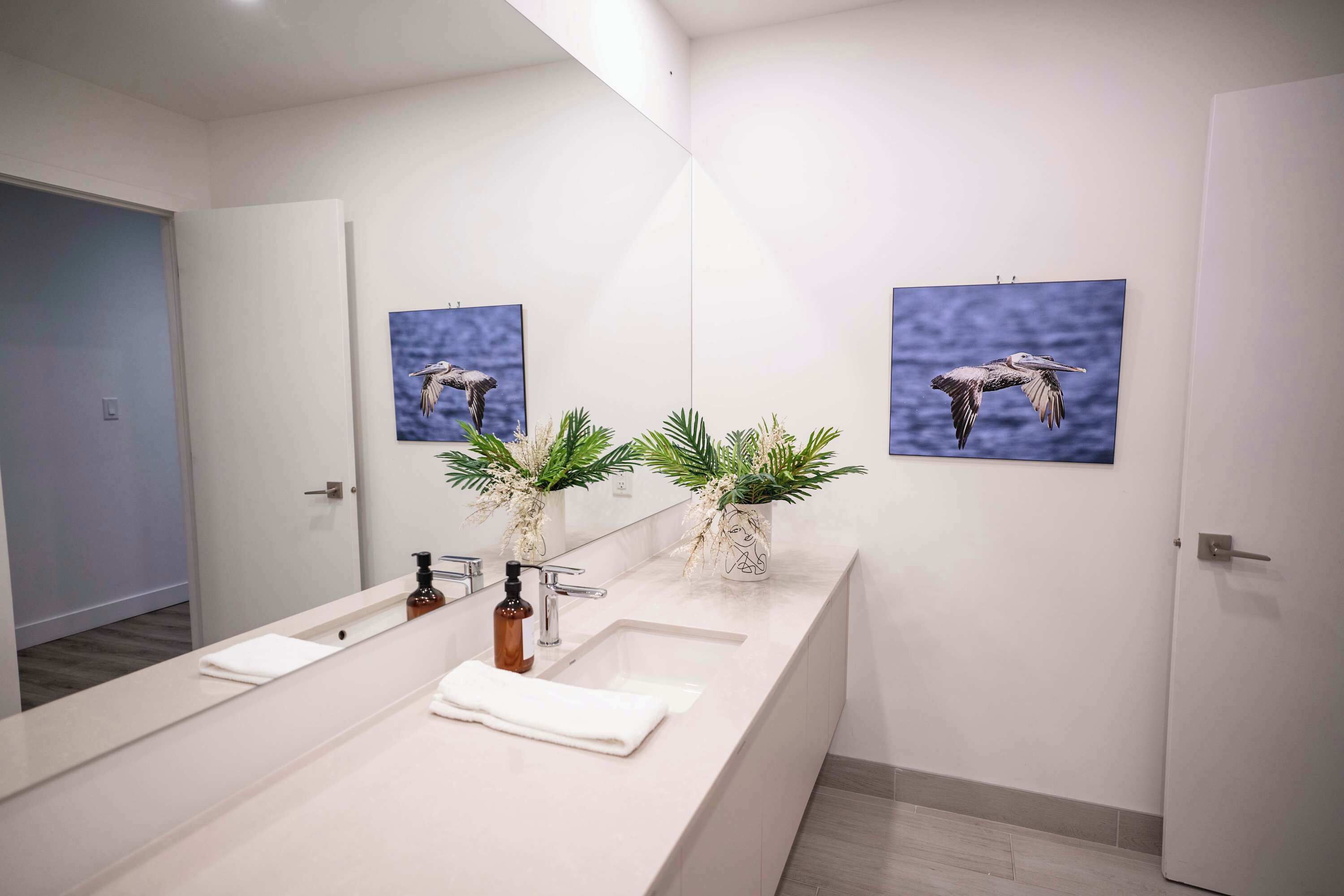 Une salle de bain moderne avec un long meuble-lavabo, deux vasques murales, un grand miroir et deux tableaux représentant des oiseaux.