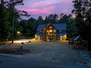 A two-story lodge with a stone facade and a large front porch, surrounded by trees and a gravel driveway. - ATP~4Kings+bunkroom~Sleeps16~Pickleball~Hottub (Broken Bow)