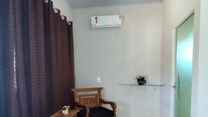 Een compacte, goed verlichte kamer met een houten bank, een houten stoel, een kleine tafel met een bloevas en een wandairconditioner.