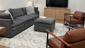A modern living room with a sectional sofa, a brown leather armchair, a grey ottoman, and a flat-screen TV on a wooden cabinet.