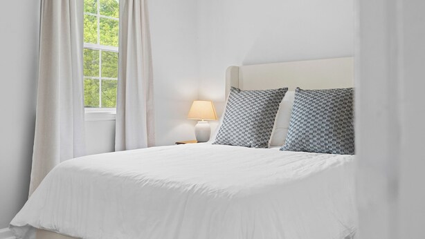 A neatly made bed with white bedding and patterned pillows, a bedside lamp, and a window with curtains.