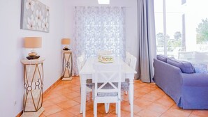 Quarto com uma mesa de jantar branca, duas cadeiras, um sofá, uma luminária e um quadro na parede.
