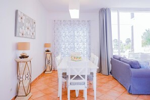 Ein Zimmer mit einem weißen Esstisch, zwei Stühlen, einer Couch, einer Lampe und einem Gemälde an der Wand.