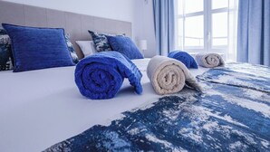 Cama bem arrumada com travesseiros azuis e brancos, edredom estampado em azul e branco, e toalhas enroladas sobre a cama.