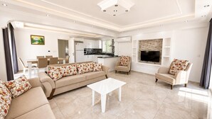 Ein modernes Wohnzimmer mit einer Couch, Sesseln, einem Couchtisch, einem Fernseher und einer Küche im Hintergrund.