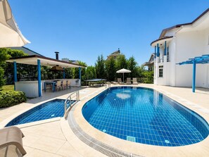 Una piscina con dos zonas diferenciadas, un área de estar techada con mesas y sillas, y un edificio de fachada blanca y detalles azules.
