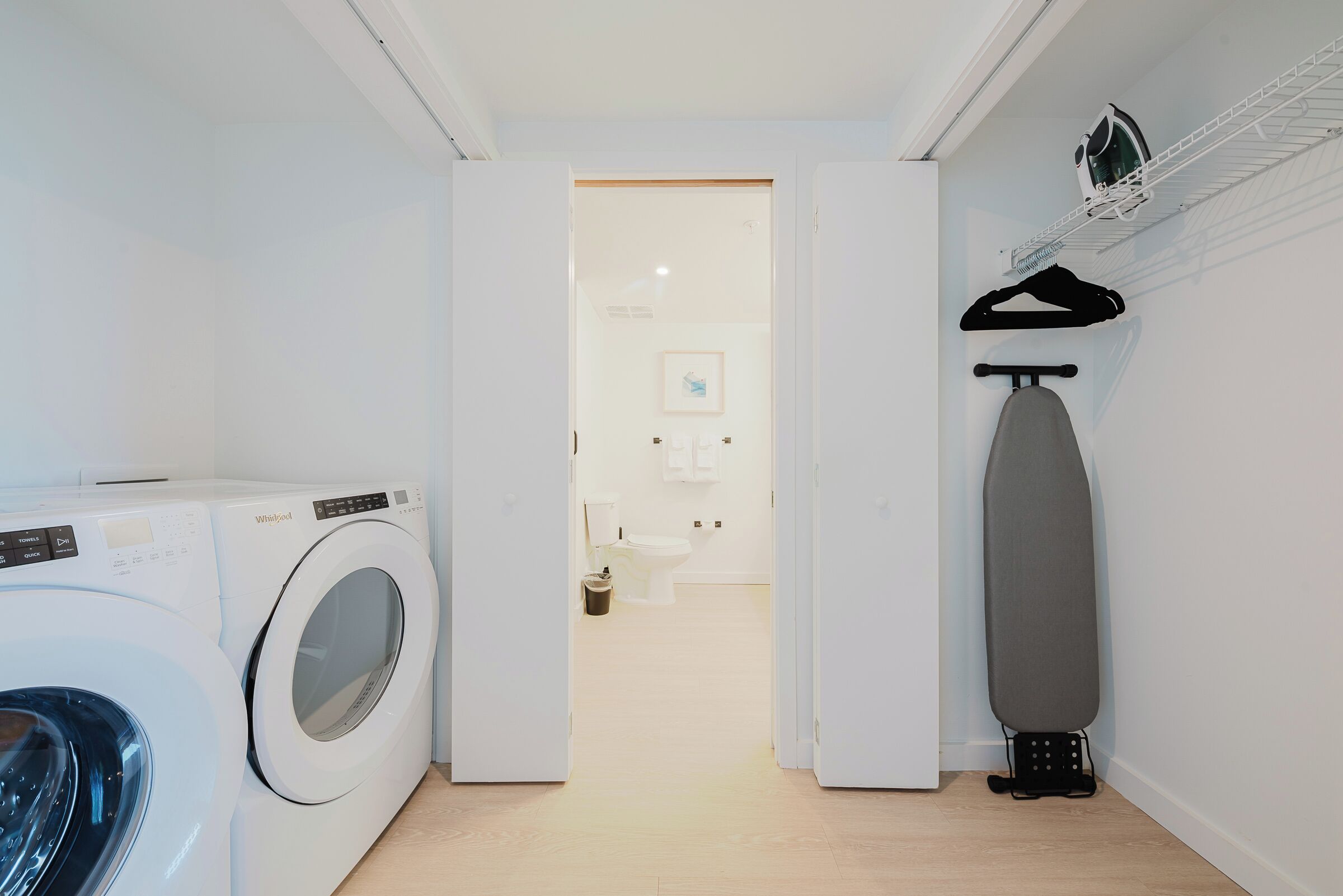 A laundry area with a front-loading washer and dryer, an ironing board, and a hanging rack.