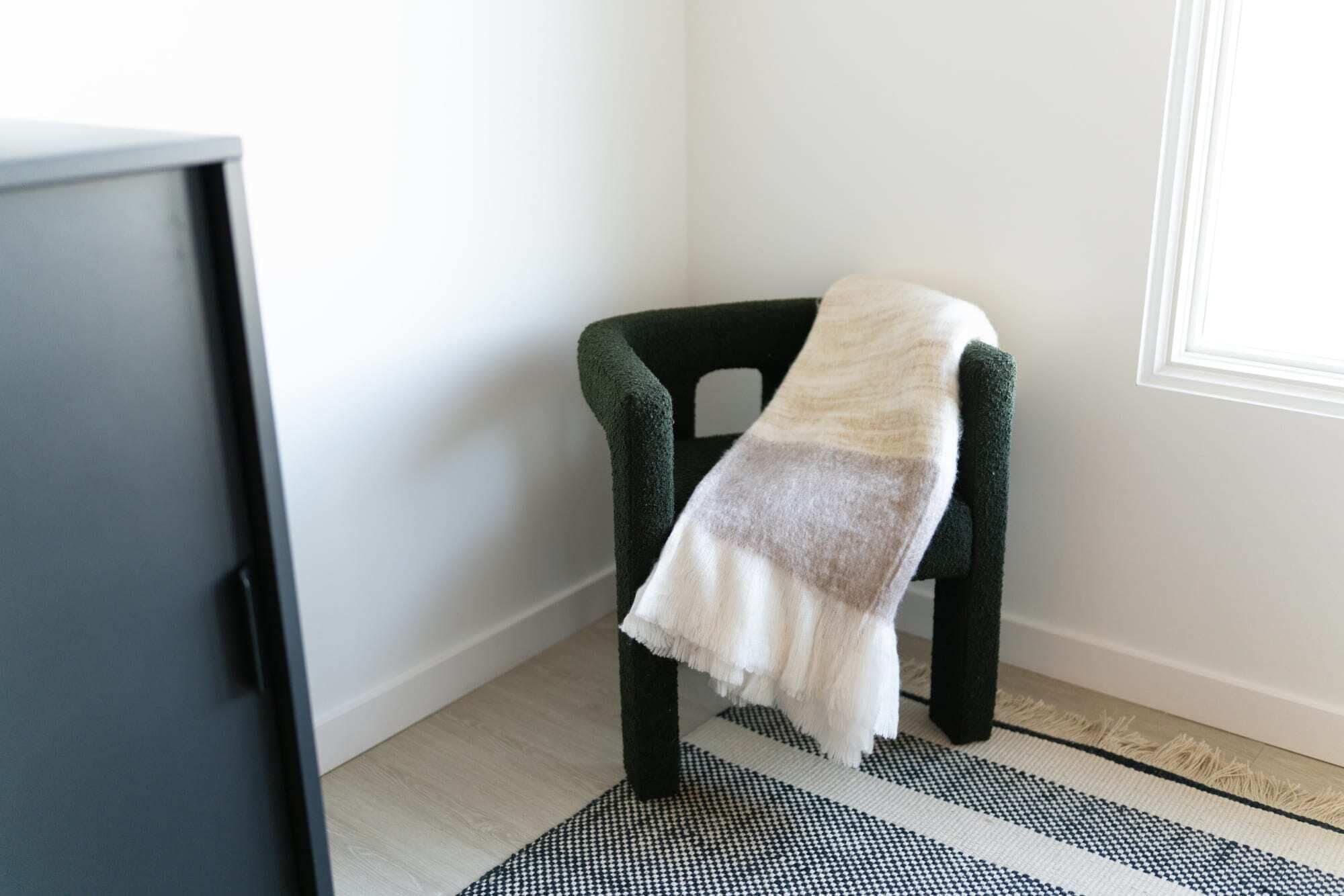 A chair with a blanket, a black and white rug, and a white wall with a window.