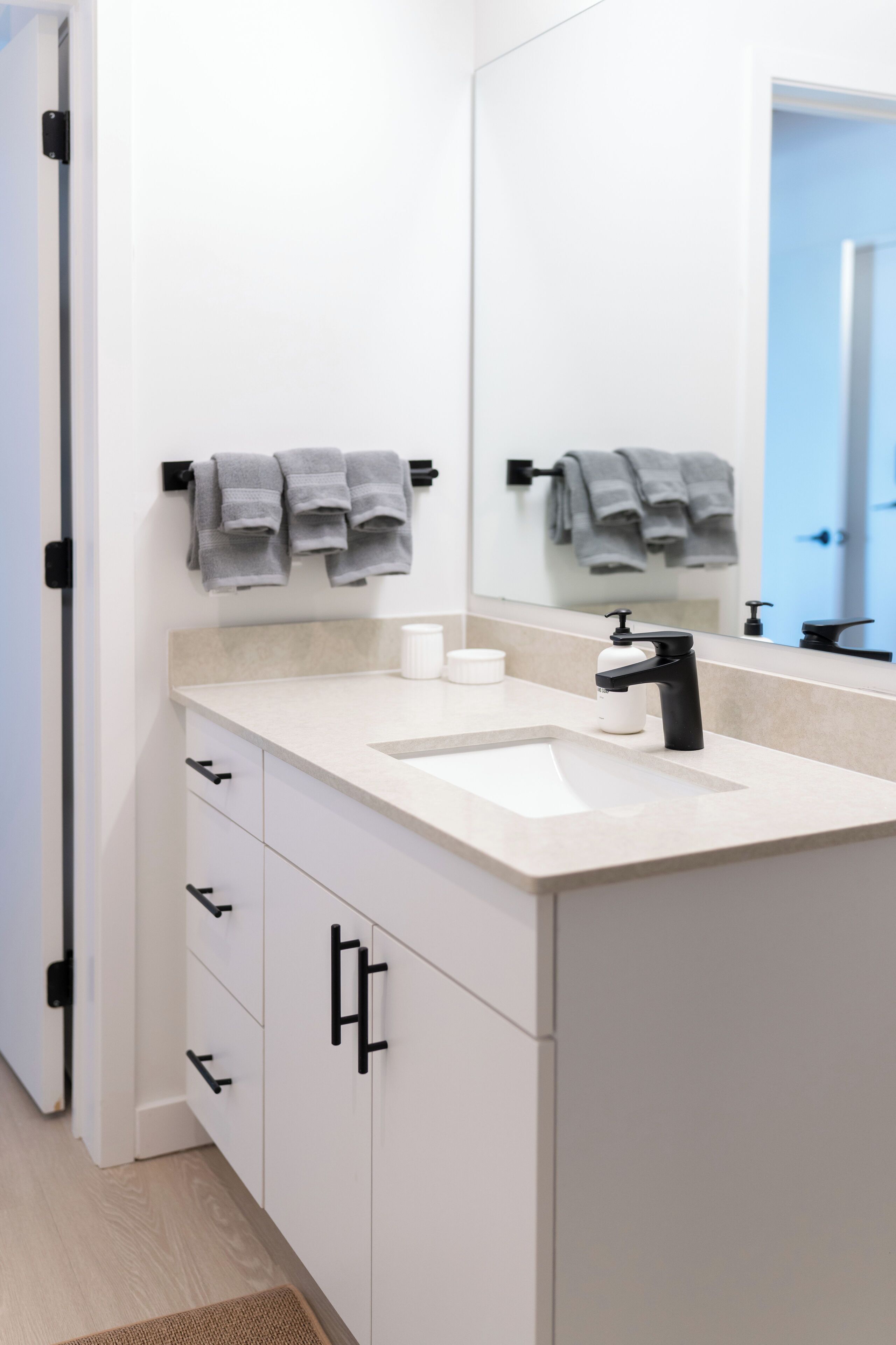 A modern bathroom with a white sink, black faucet, and a mirror above it. There are towels hanging on a rack and a small white container on the counter.