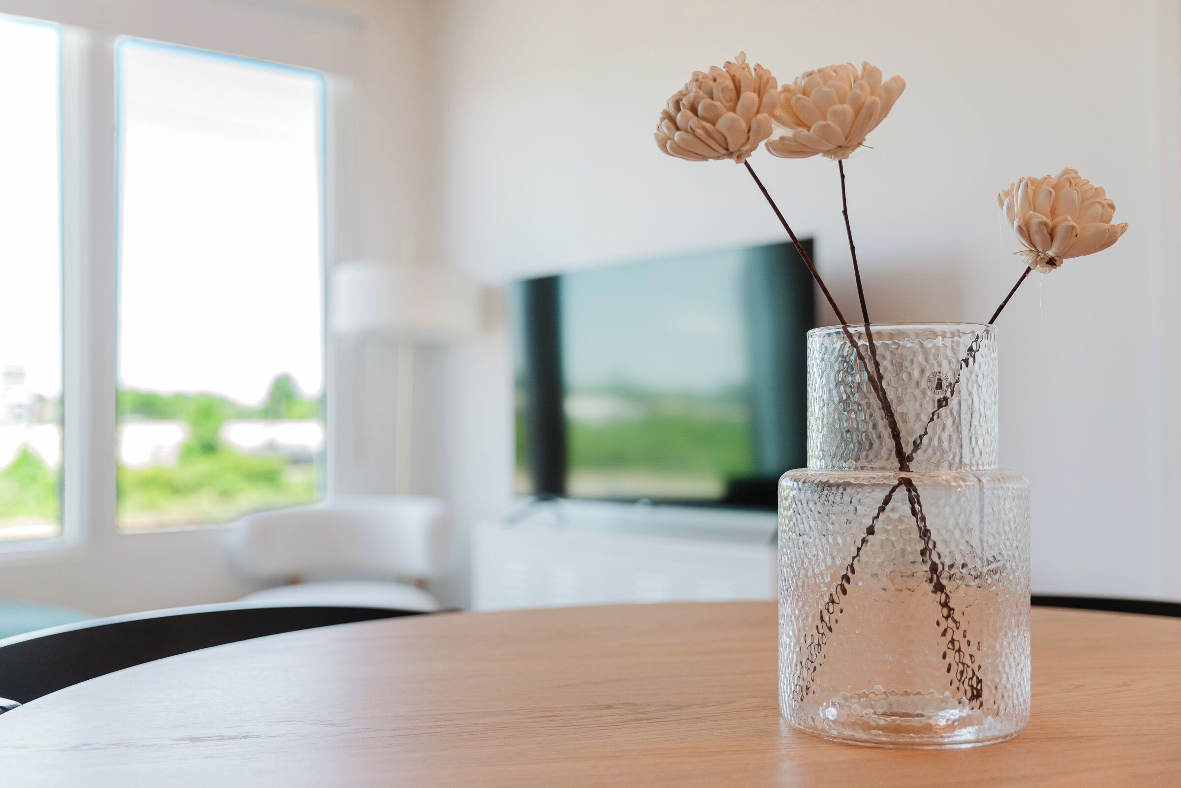 Un vase en verre transparent, orné de fleurs, est posé sur une table en bois dans une pièce où se trouvent une télévision et une fenêtre.