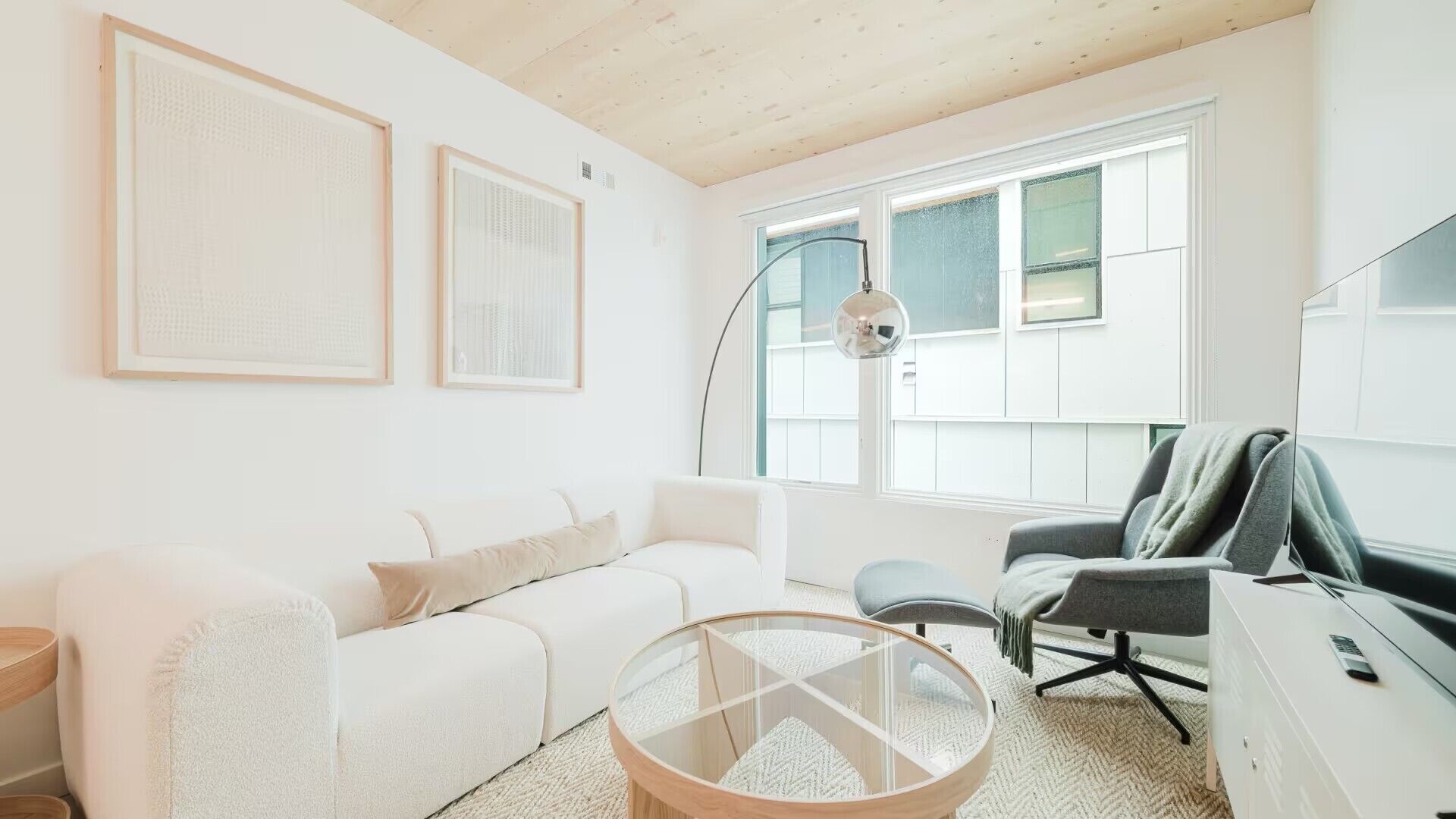 Uma sala de estar moderna com um sofá branco, uma mesa de centro de vidro, uma cadeira de escritório cinza e uma escrivaninha com um computador.
