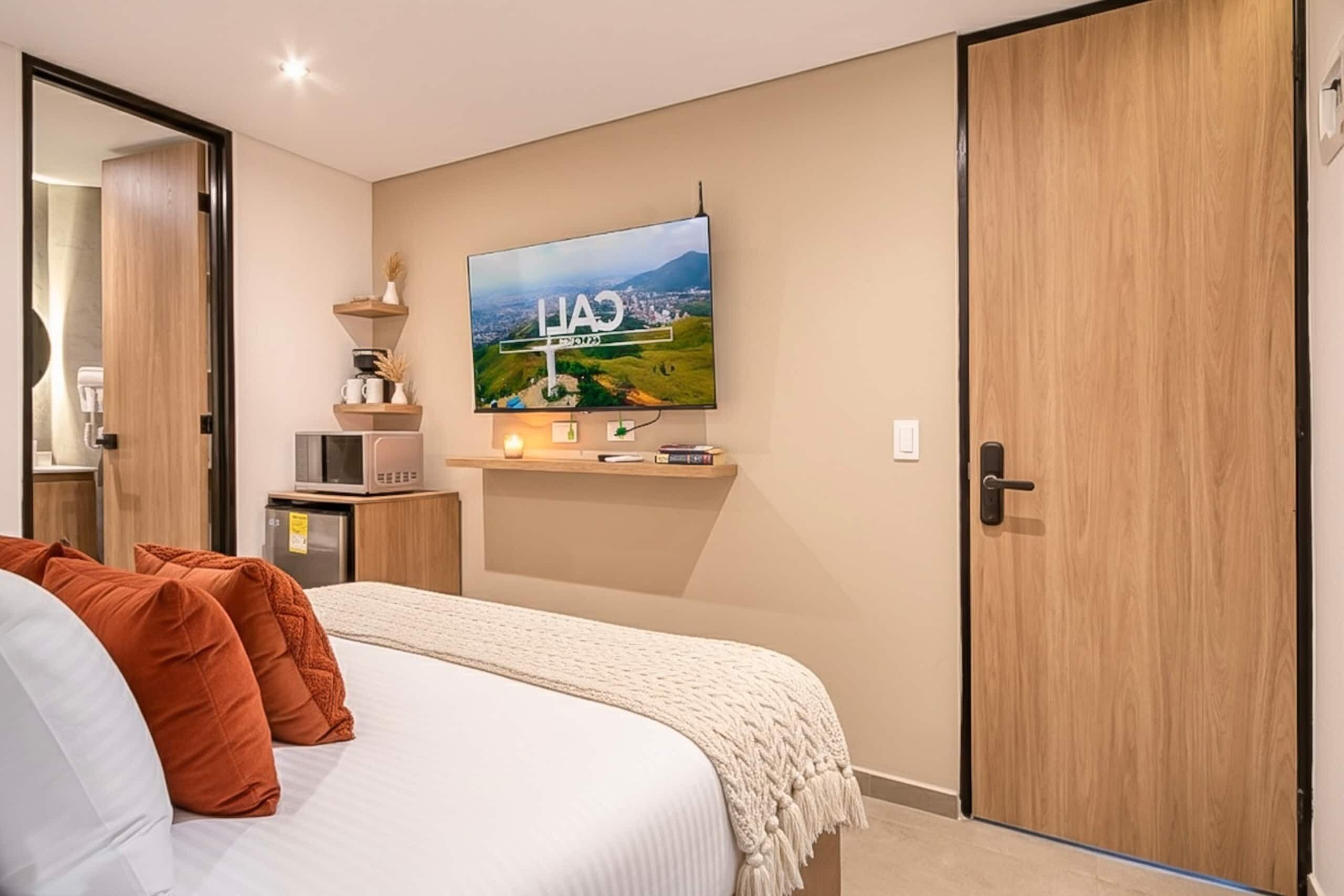 Un dormitorio con una cama, un televisor montado en la pared, un estante con objetos y una puerta.
