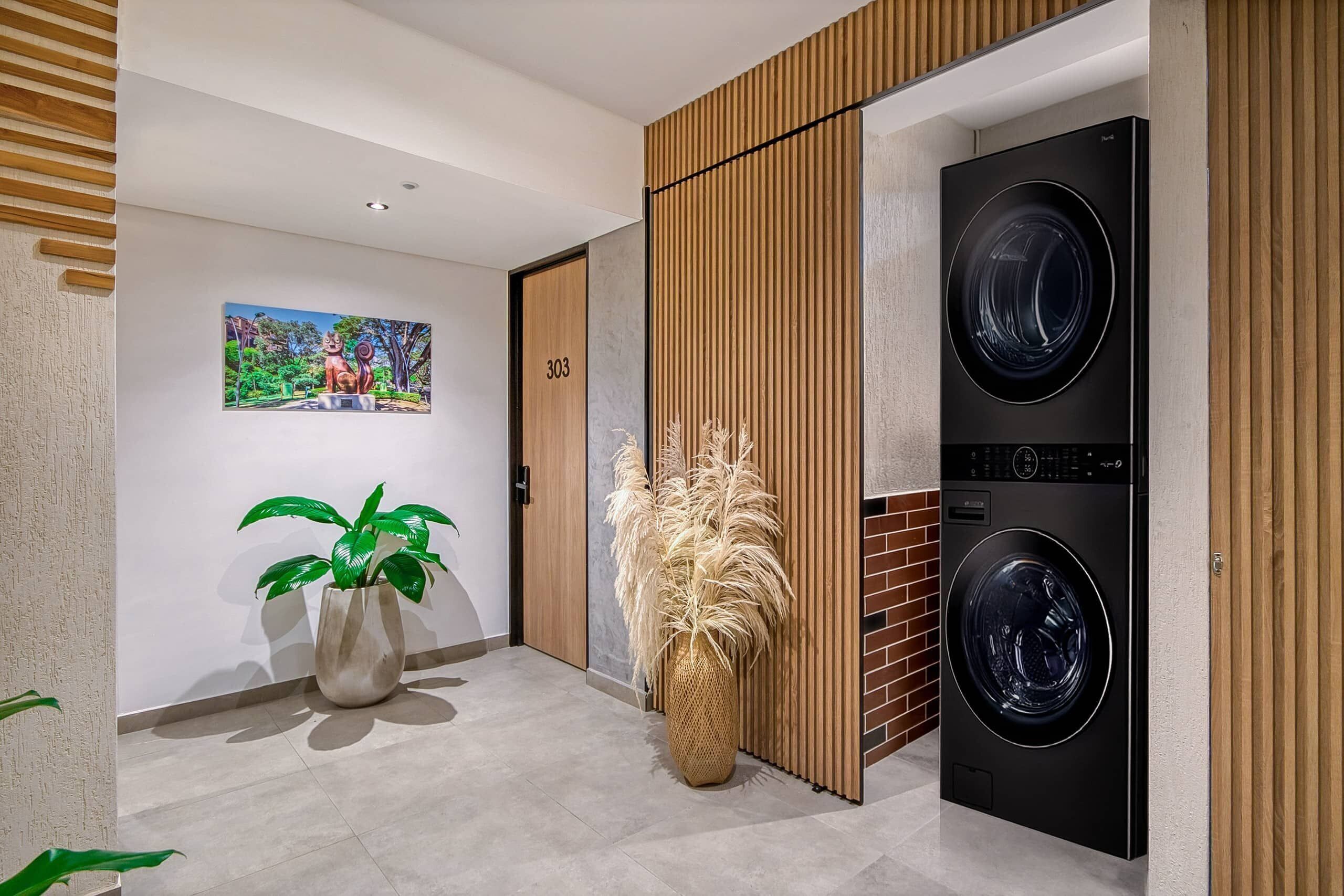A modern laundry room with two black washing machines, a wooden panel wall, a potted plant, and a framed picture on the wall.