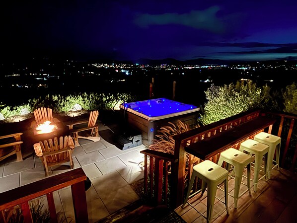 Un patio al aire libre con jacuzzi, asientos y vista a un paisaje urbano nocturno.