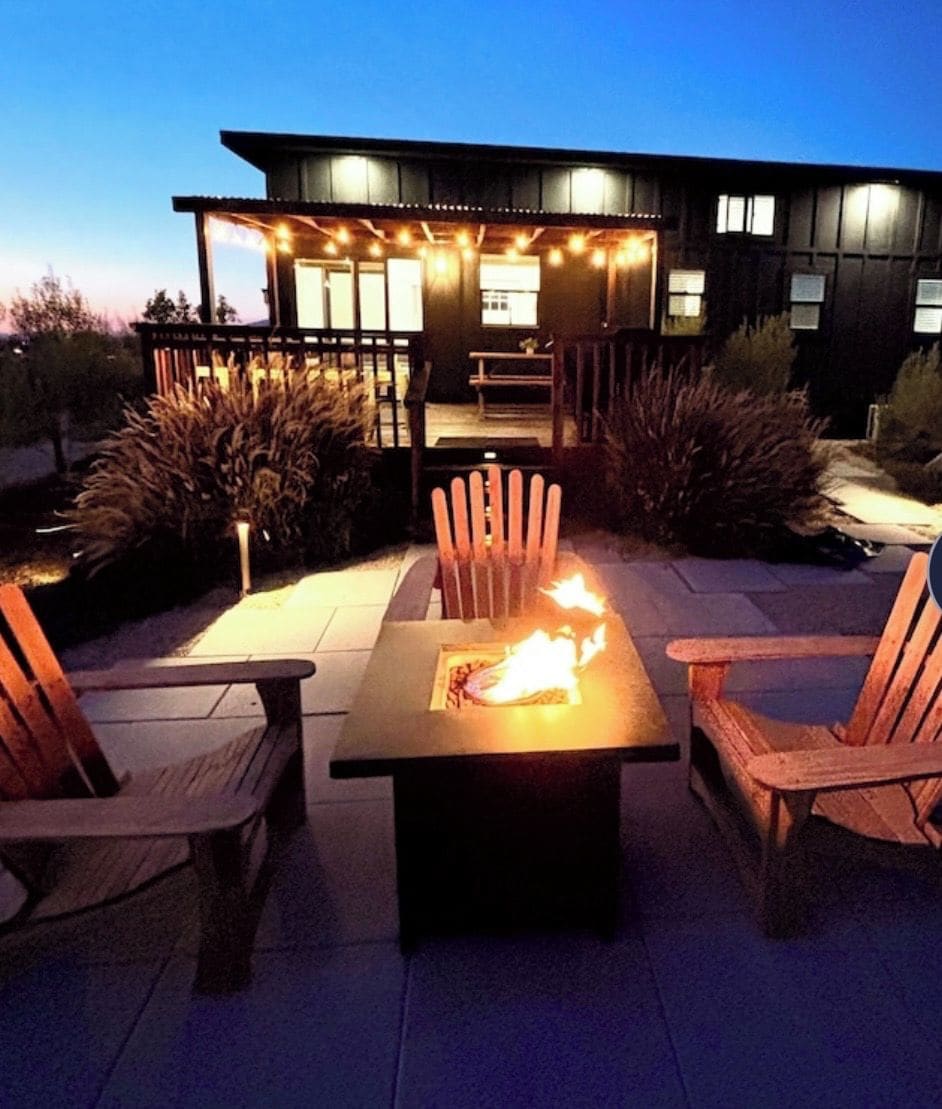 An outdoor patio with a fire table and chairs, surrounded by plants and a modern building in the background.