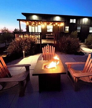 Une terrasse extérieure avec une table et des chaises autour d’un foyer, entourée de plantes et avec un bâtiment moderne en arrière-plan.