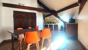 Un coin repas avec une table en bois et des chaises orange, une cuisine en arrière-plan et un plafond en pente avec des poutres apparentes.