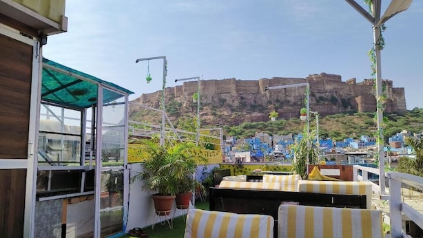 Terraço no telhado com vista para uma fortaleza histórica, área de estar com almofadas listradas e plantas em vasos.