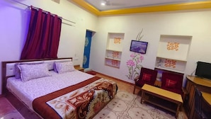 Quarto de hotel com cama, escrivaninha, cadeira, televisão e prateleiras decorativas na parede.