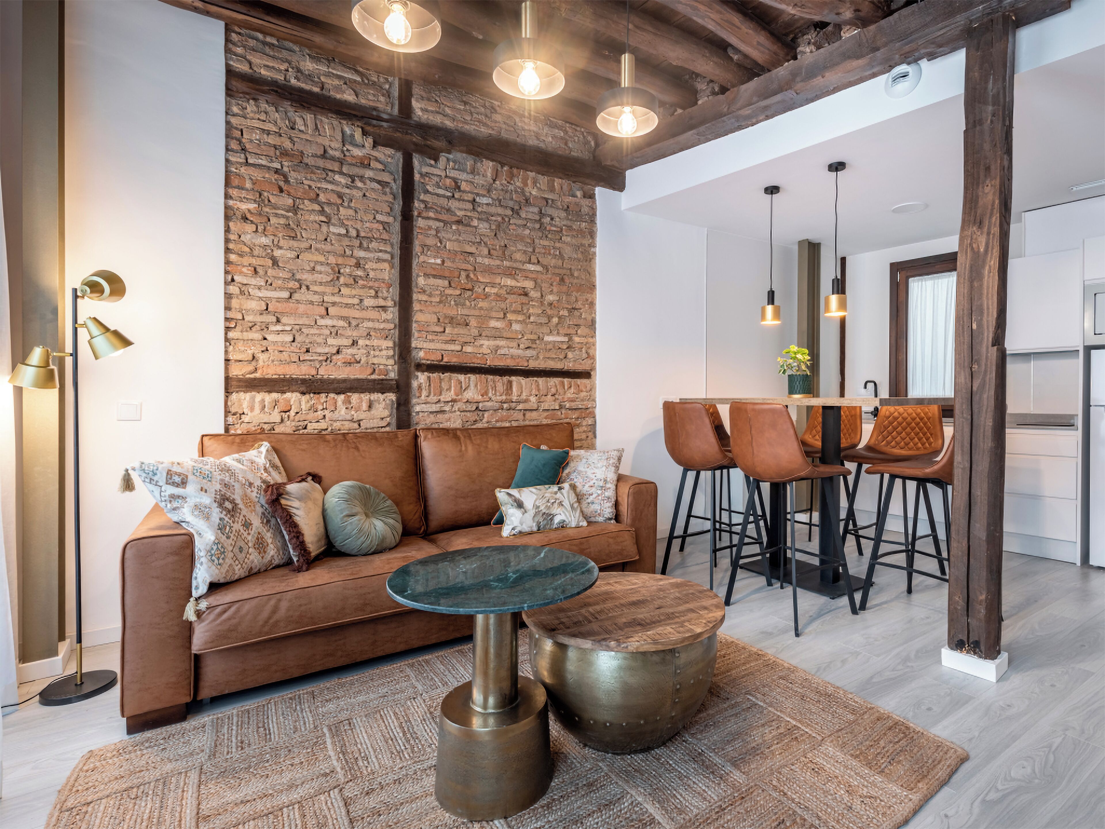 Una sala de estar moderna con un sofá de cuero marrón, una mesa de centro redonda y una pared de ladrillo. Se observan dos taburetes y una zona de cocina al fondo.
