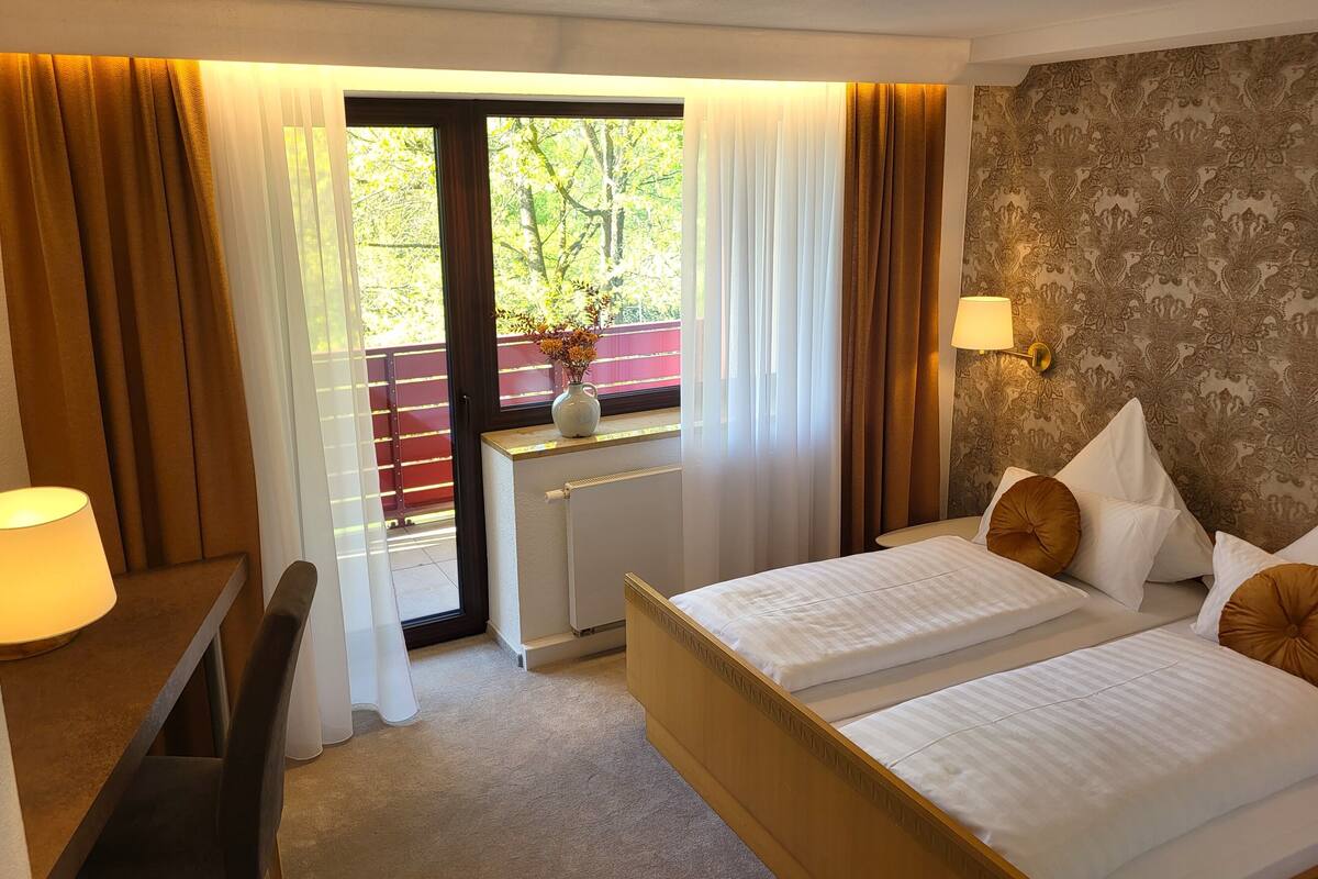 A hotel room with two beds, a desk with a lamp, a window with curtains, and a radiator.