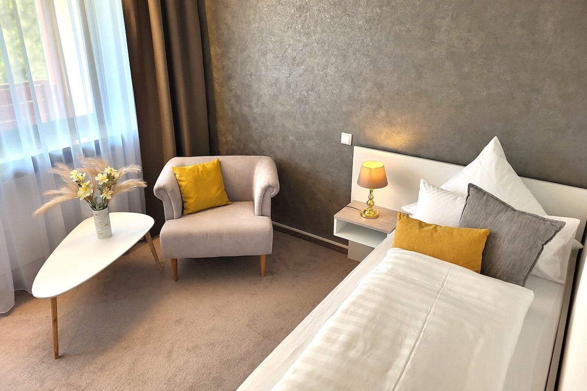 A hotel room with a bed, a chair, a nightstand with a lamp, a small table with a vase of flowers, and a view through sheer curtains.