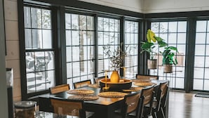 Zona pranzo con tavolo e sedie in legno, ampie finestre e piante pensili.