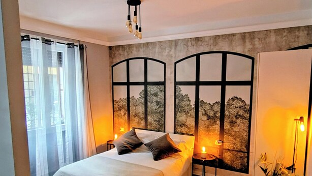 A modern bedroom with a large bed, two bedside tables with lamps, a view of trees through a window, and a wardrobe.