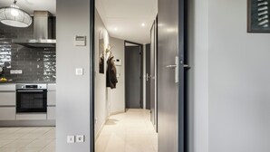 A modern kitchen with a built-in oven, a stainless steel range hood, and a pendant light. A hallway leads to a door with a mirror and a coat rack. - Vivre Cannes (Cannes)