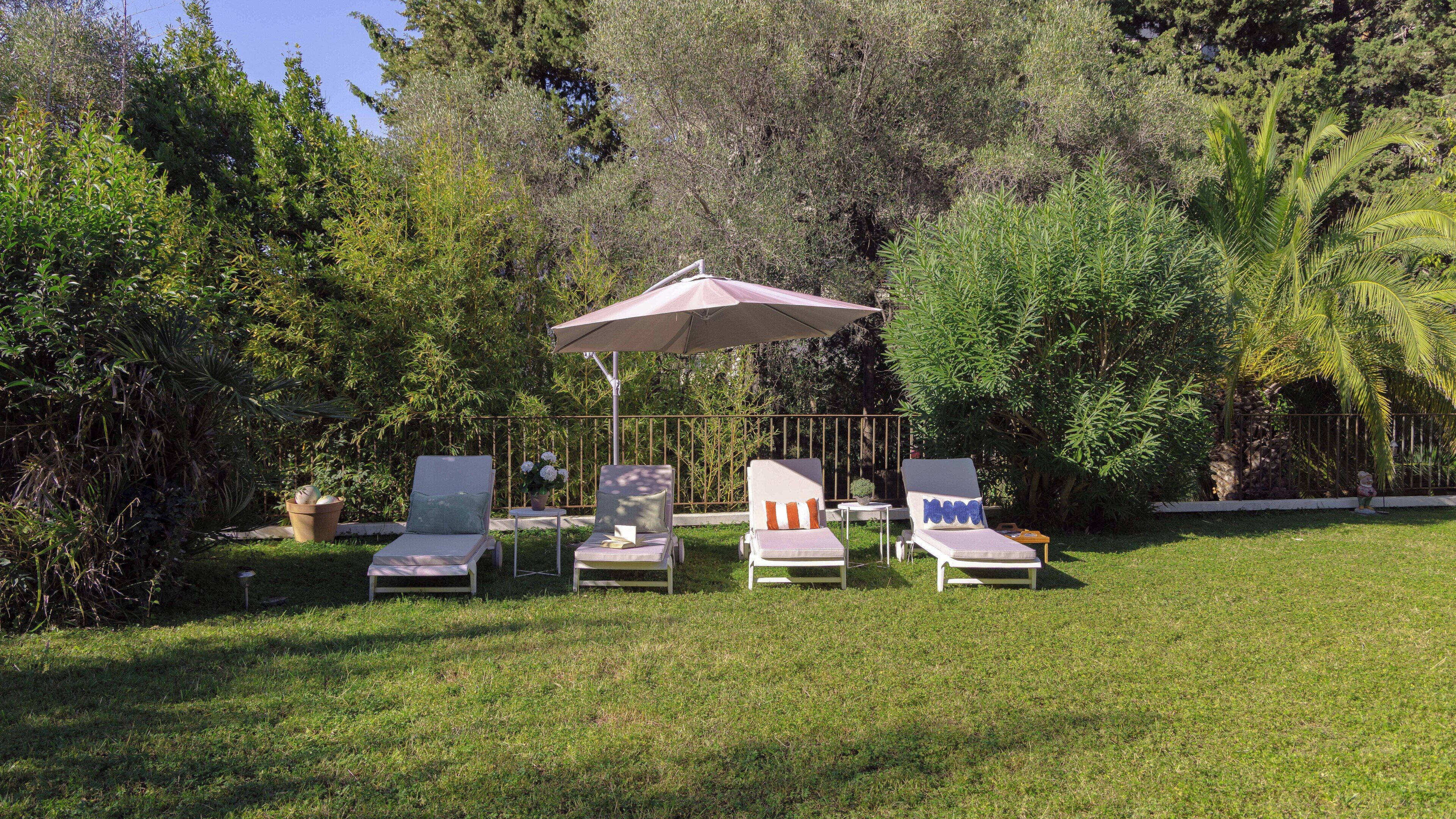 A garden with four lounge chairs under a large umbrella, surrounded by lush greenery.