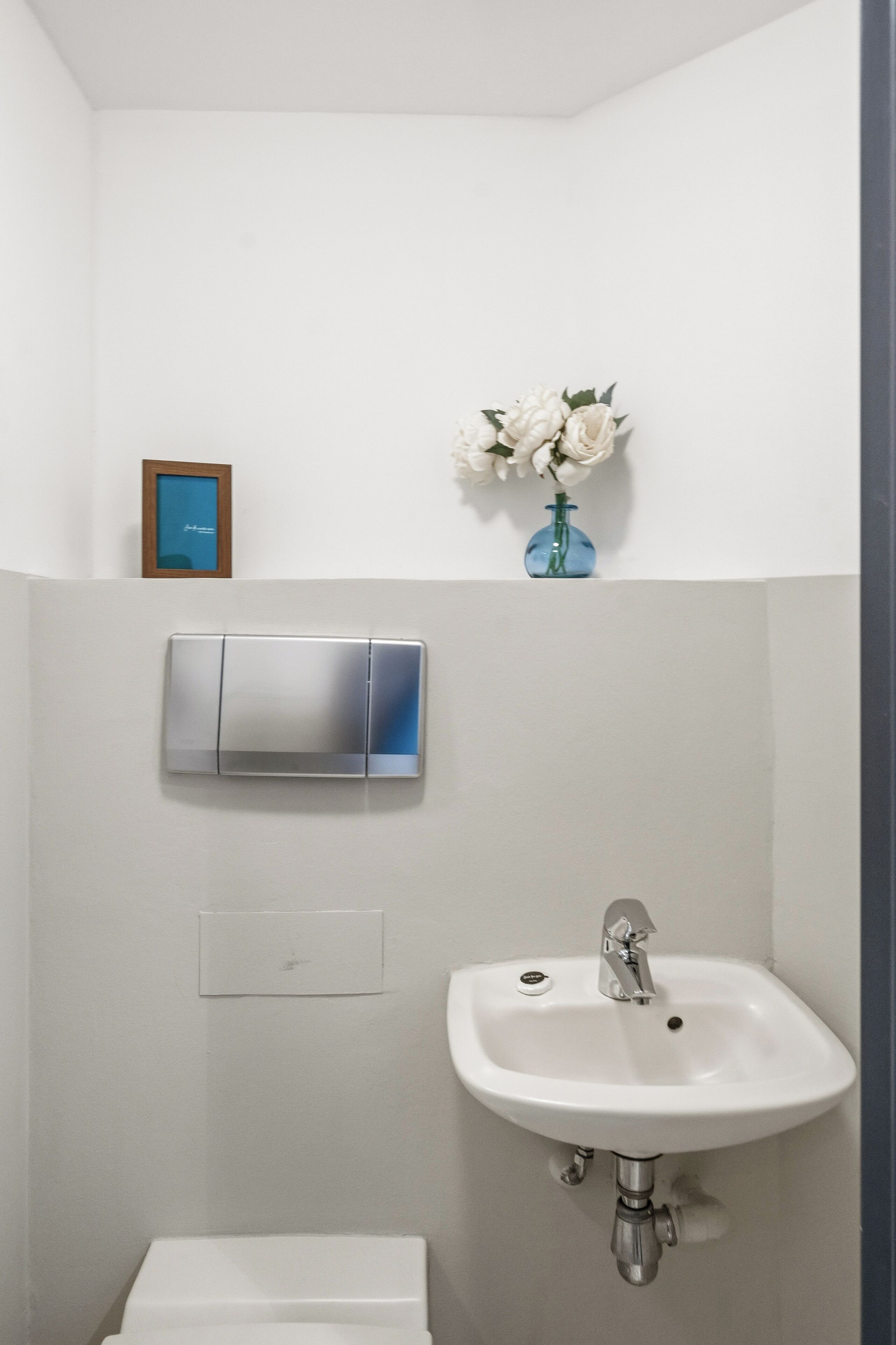 Un baño con un lavamanos blanco, un espejo y un pequeño jarrón con flores en la pared.