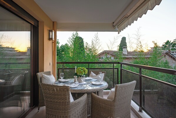 A balcony with wicker furniture, a table set for two, and a view of greenery and buildings in the background.