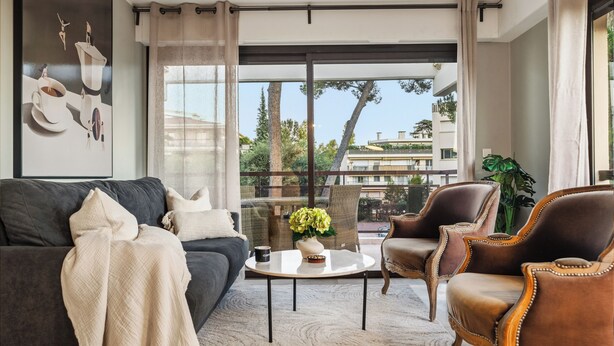 A modern living room with a dark sofa, a round coffee table, and two brown armchairs. A large window offers a view of trees and buildings outside. - Vivre Cannes (Cannes)