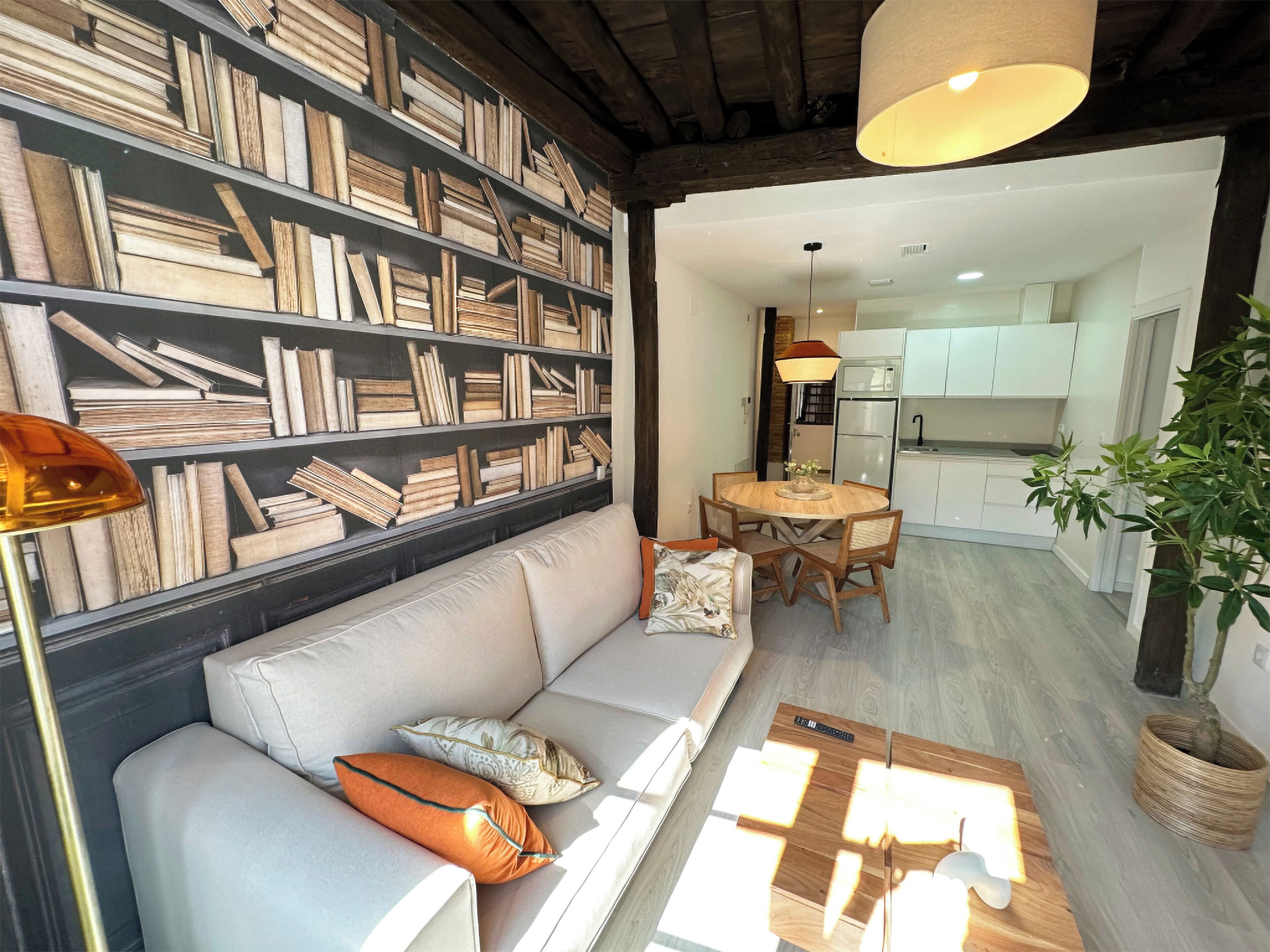 A living room with a sofa, a coffee table, and a bookshelf filled with books. There is a dining area with a table and chairs, and a kitchen in the background.