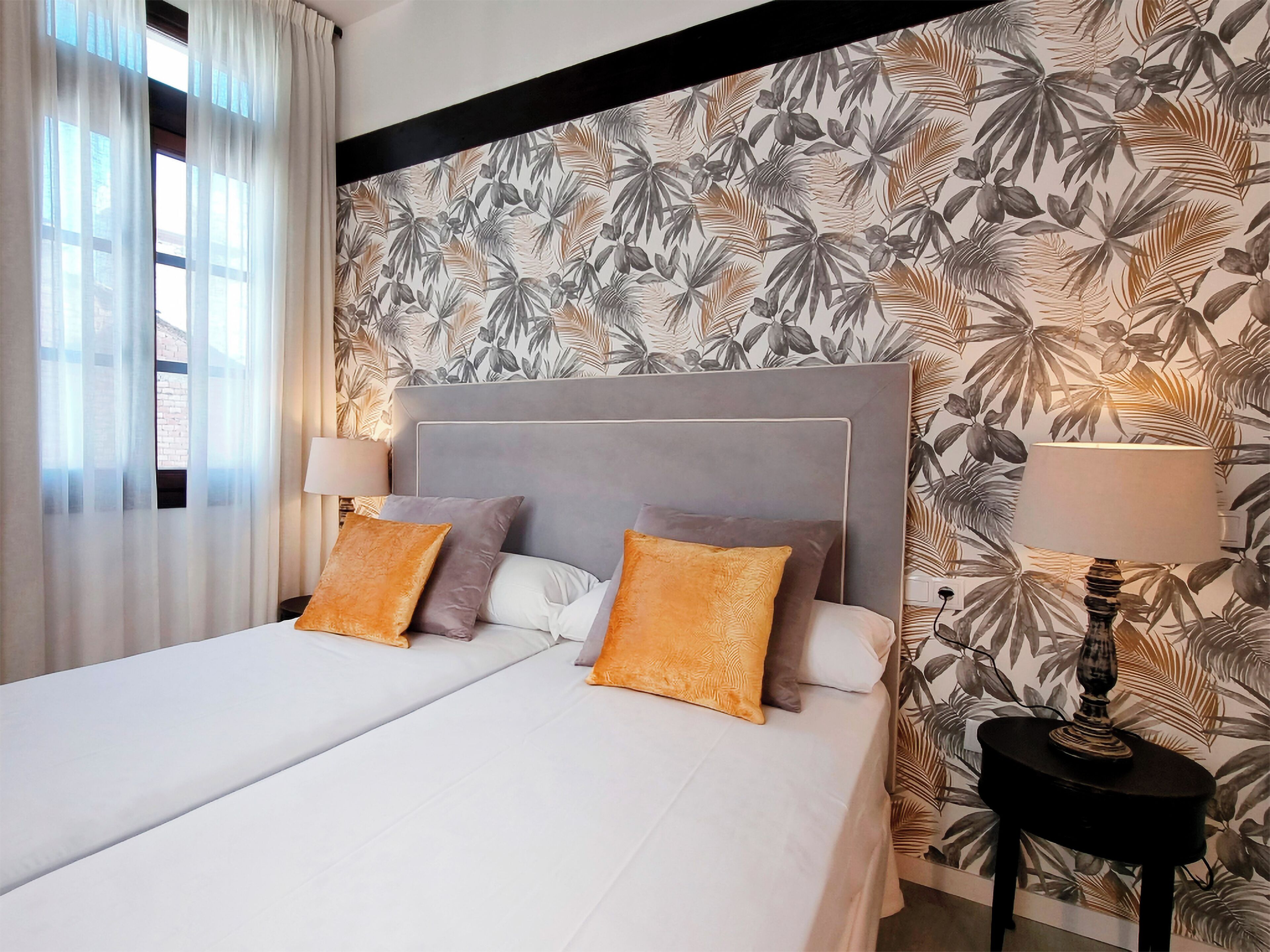A hotel room with a large bed, two pillows, a nightstand with a lamp, and a wall with a tropical leaf pattern.