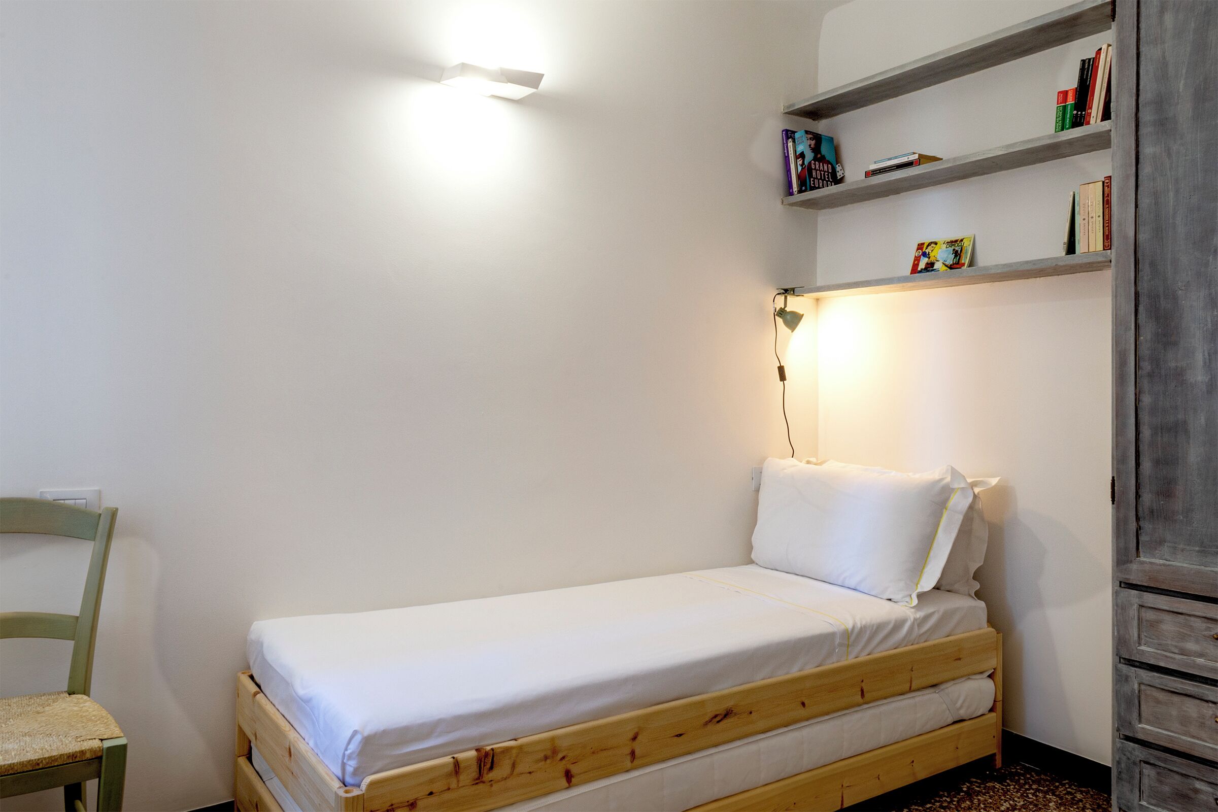 A small, single-bed room with a wooden frame, a green chair, a bedside table, and a shelf with books.