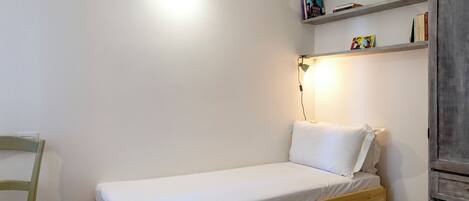 A small, single-bed room with a wooden frame, a green chair, a bedside table, and a shelf with books.