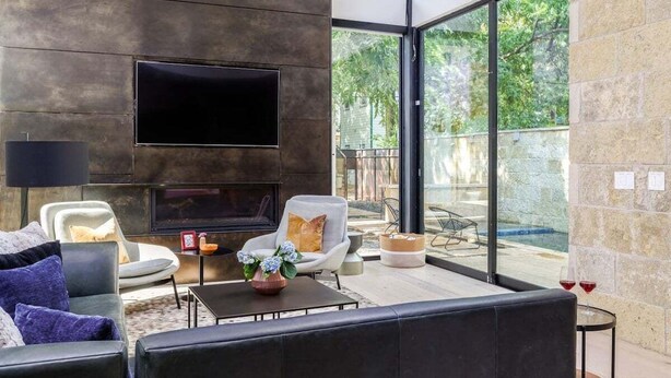 Een moderne woonkamer met een flatscreen-televisie, zachte zitplaatsen, een bijzettafeltje en uitzicht op een zwembad en groen.