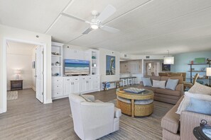 A spacious living area with a ceiling fan, a television, a dining table, and a kitchen area in the background. - Sandcastle 401 (New Smyrna Beach)