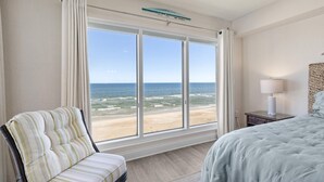 Une chambre avec une grande fenêtre donnant sur la plage, un lit, une chaise et une table de chevet avec une lampe.