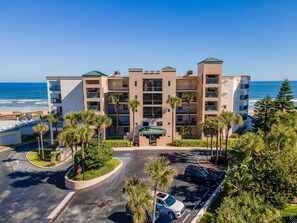 A multi-story hotel with a parking lot, palm trees, and a view of the ocean. - Sandcastle 401 (New Smyrna Beach)