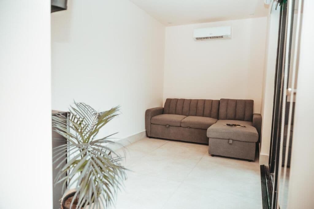 Ein Wohnzimmer mit einer Couch, einem Hocker, einer Topfpflanze und einer Klimaanlage an der Wand.